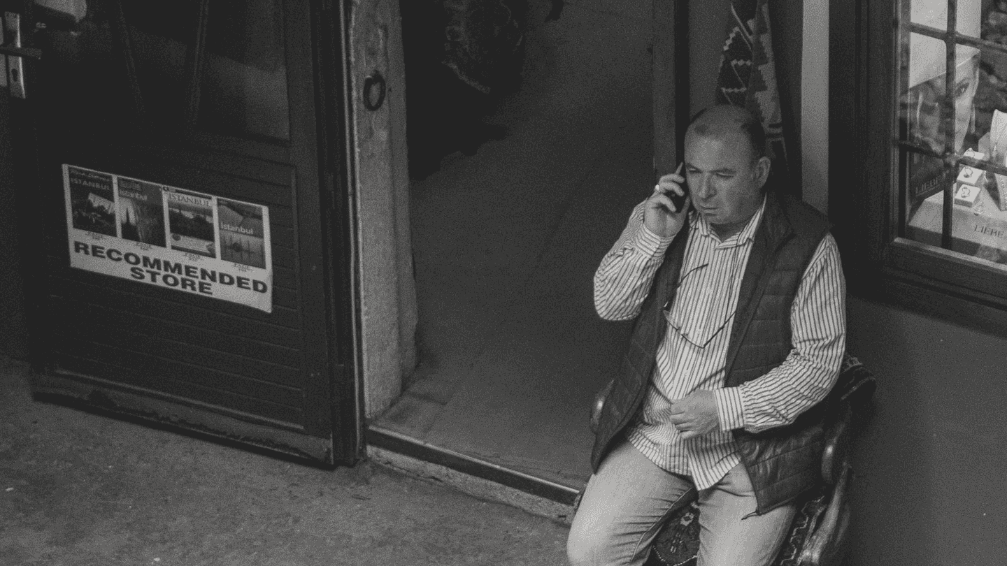 A man on the phone outside a store, sitting against the front of it.